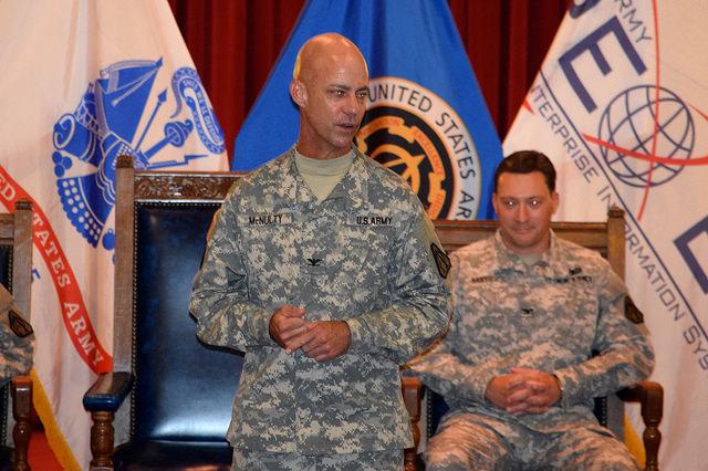 Soldiers addresses crowd while another soldier sits in a chair behind him.