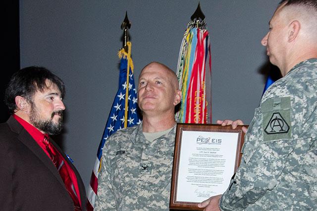 Three men stand on stage in front of flags. Man on far right hands a framed piece of paper to man on left.