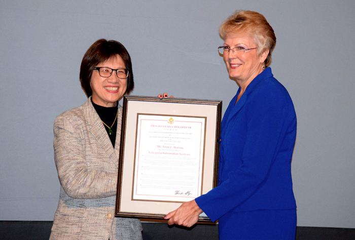 Two women stand next to one another. They hold a framed certificate together.